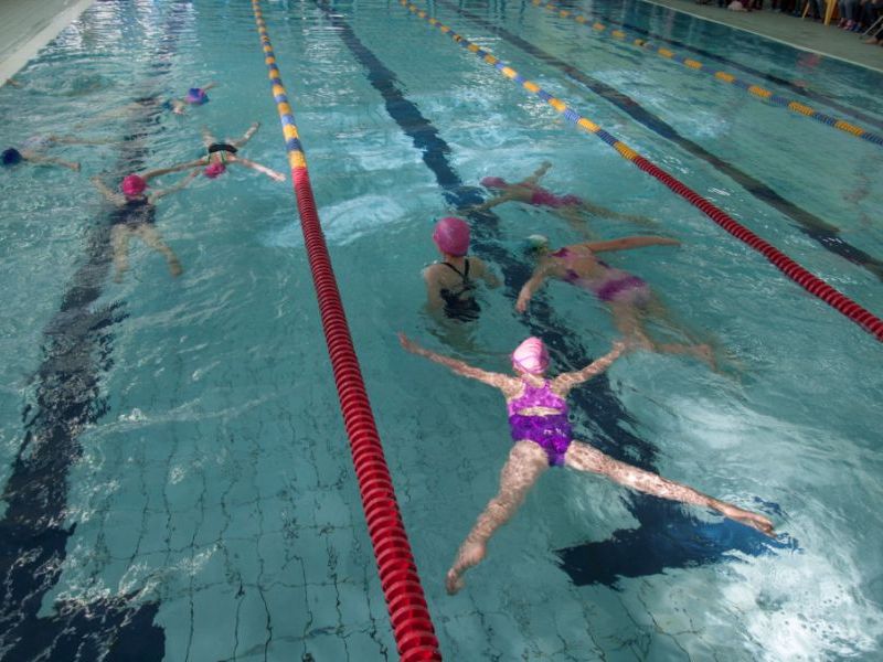 Na zdjęciu dzieci pływające na torze basenu w pełnym zanurzeniu. Na głowach różowe czepki, różowe kostiumy kapielowe