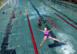 Na zdjęciu dzieci pływające na torze basenu w pełnym zanurzeniu. Na głowach różowe czepki, różowe kostiumy kapielowe